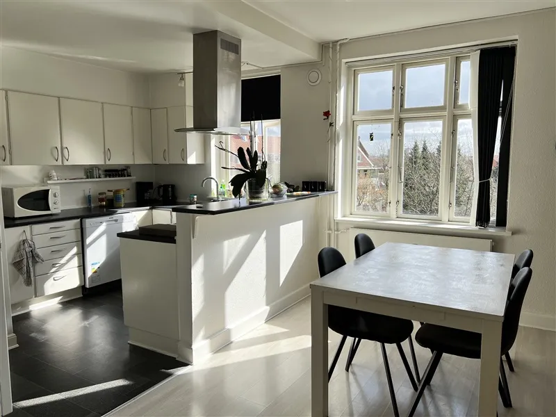 Moderne køkken med hvide skabe, spisestue med borde og stole og udsigt til træer og huse fra vinduet