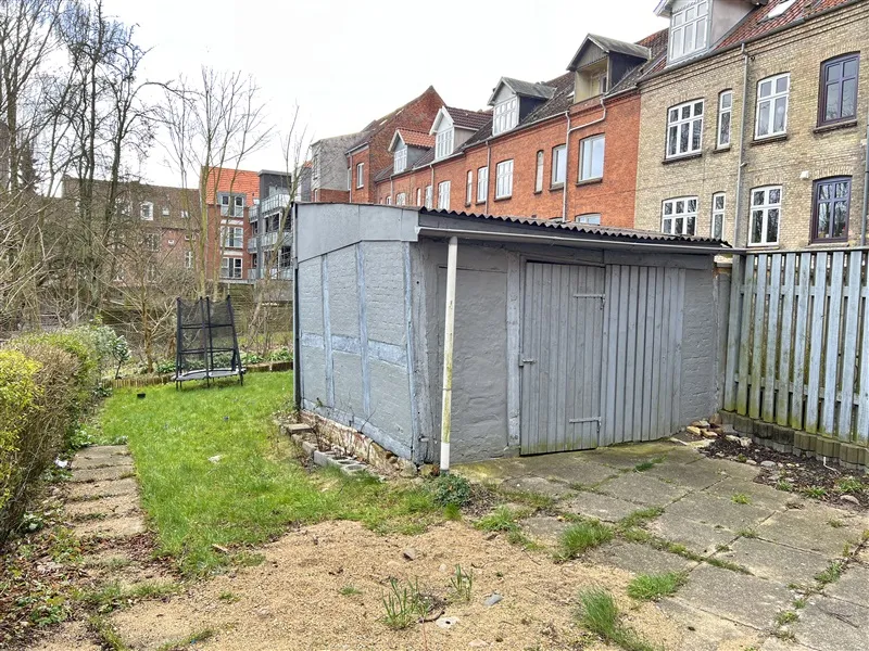 Grå værkstedsbygning med hæk og grønne græsflæder ved siden af en høj mur og røde bygninger i baggrunden