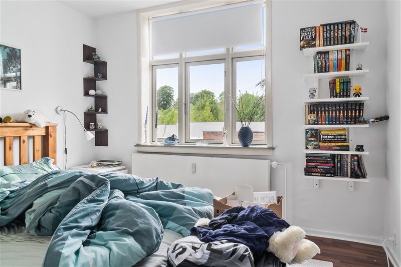 Et soveværelse med hvidt loft og vægge. Et enkelt enkeltseng med lyse blå tæppe og skumhovedpude. Et skrivebord med bøger og