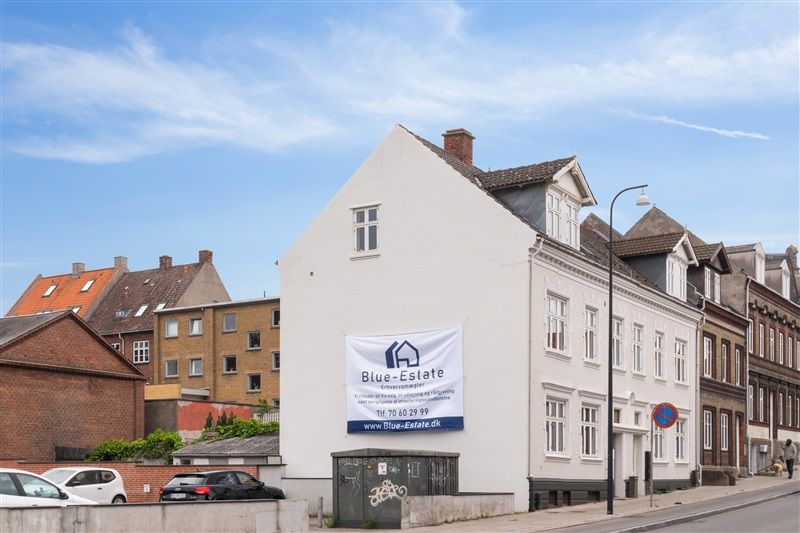 En hvid hus med et banner med Blue Estate tekst og kontaktoplysninger er placeret i en by med biler parkeret og en person