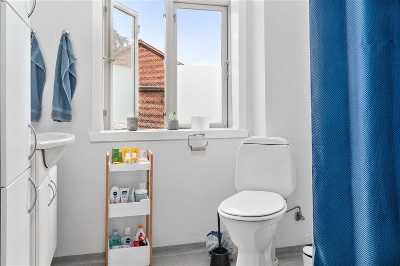 Toilet med toiletpapirholder, toiletbørste og urinal. Toilet med vinduesruder og toiletpapir. Rum med håndklæder og