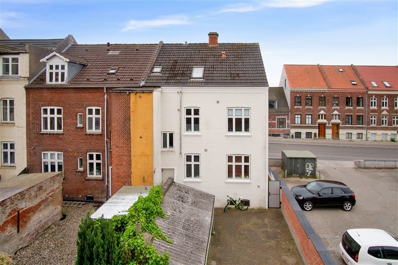 To biler parkeret i forgrunden, en mørk SUV og en hvid familiebil, parkeringsplads med grønne planter og en cykel, bagved