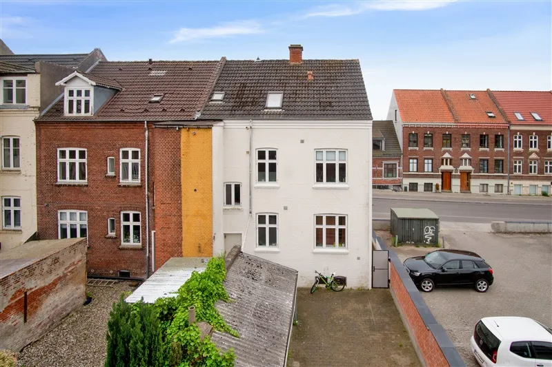 To biler parkeret i forgrunden, en mørk SUV og en hvid familiebil, parkeringsplads med grønne planter og en cykel, bagved