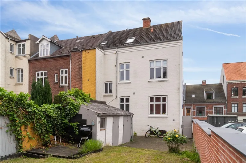 Rækkehus med grill, trælilje og to cykler i baghaven. Foroven ser man huse med røde tegl og hvide facader.