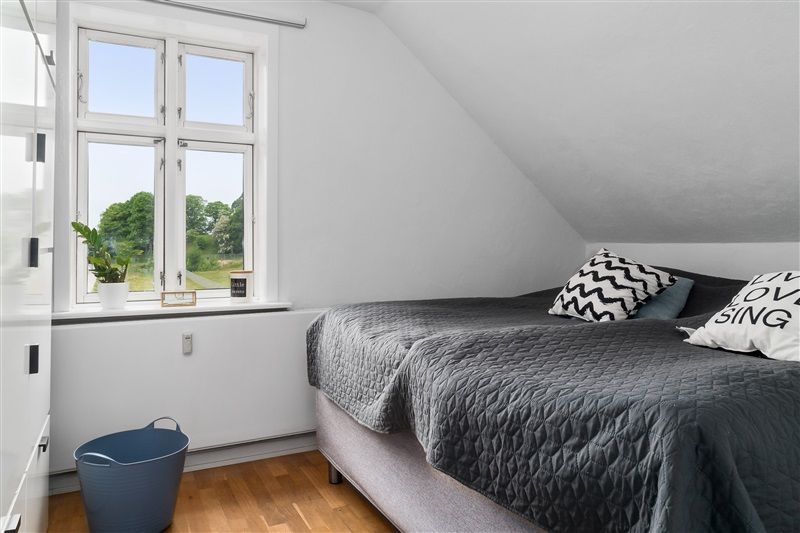 En lille soveværelse med lysegul væg, grøn blomst på vindueskarmen og et enkelt dobbeltseng med grå sengetøj og kuglepuder