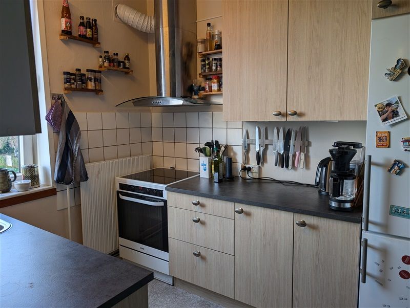 Et køkken med lyse træskabe, en hvid køleskab og en sort bordplade, en ovn og en kogeplade samt flere udstyr som knive,