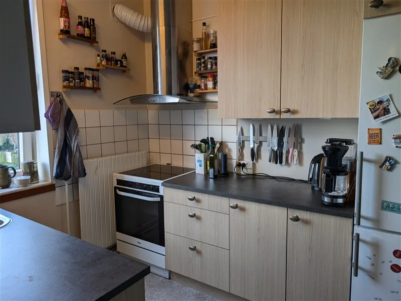 Et køkken med lyse træskabe, en hvid køleskab og en sort bordplade, en ovn og en kogeplade samt flere udstyr som knive,