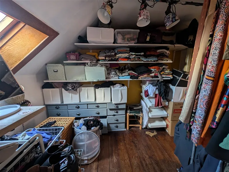 En lukket kammer med trægulv og højloft, fyld af kufferter, tøj og tøjvask. Lysere gulve og lysekroner hængende fra loftet