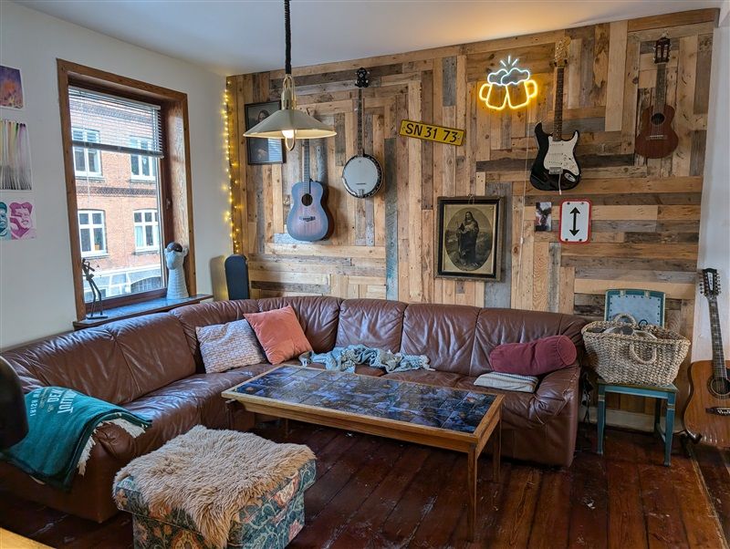 En stor brun sofa med pude og tæppe samt et massivt træbord med en blæsende lampeskærm og musikinstrumenter i baggrunden