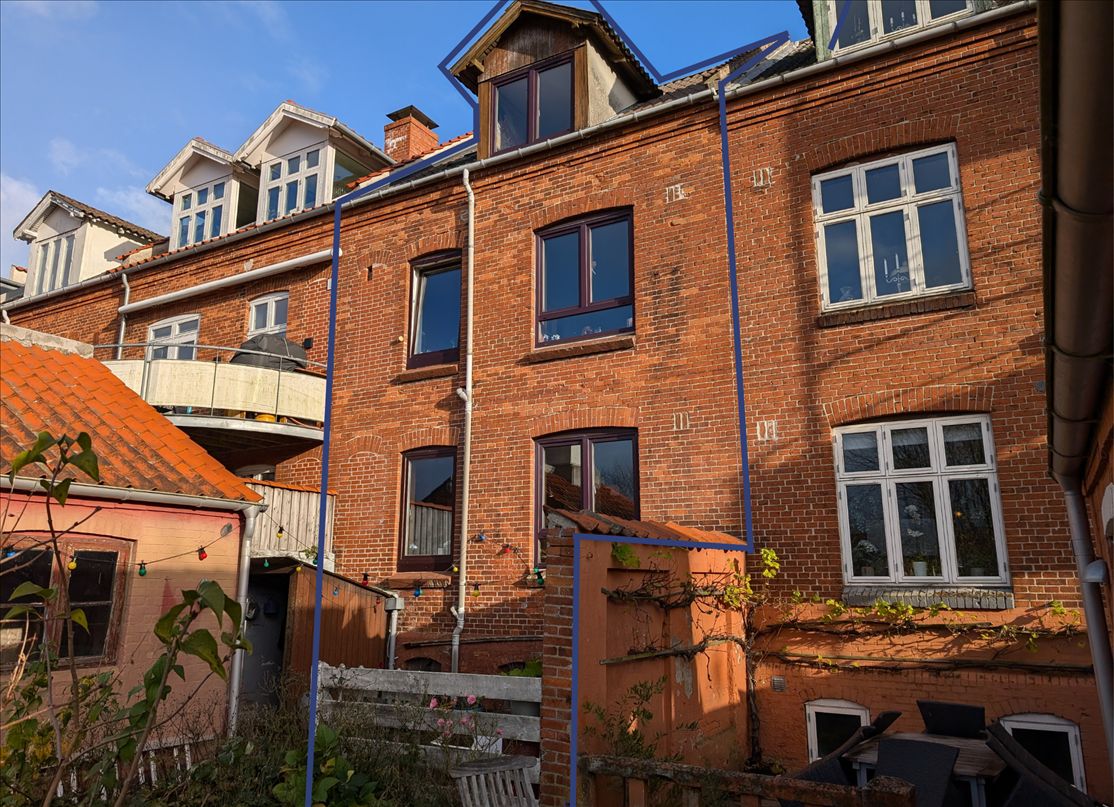 Lejlighedsbygning med røde mursten og mange vinduer, udsmykket med lysekroner og vinranker, og en privat have med møbler