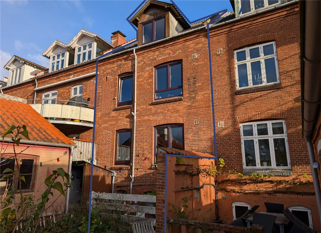 Lejlighedsbygning med røde mursten og mange vinduer, udsmykket med lysekroner og vinranker, og en privat have med møbler