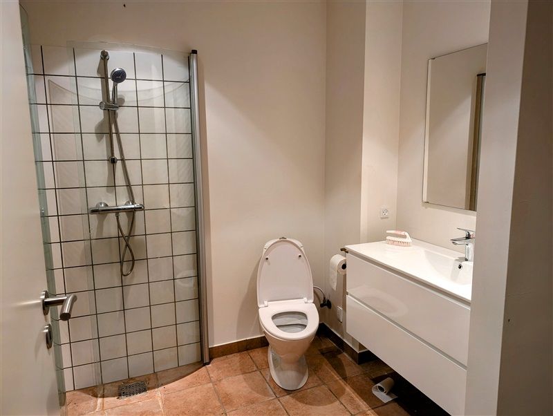 Et lille moderne badeværelse med hvidt toilet, hvidt badekar og hvidt spejl samt brusekabine med glasdør og håndbruser