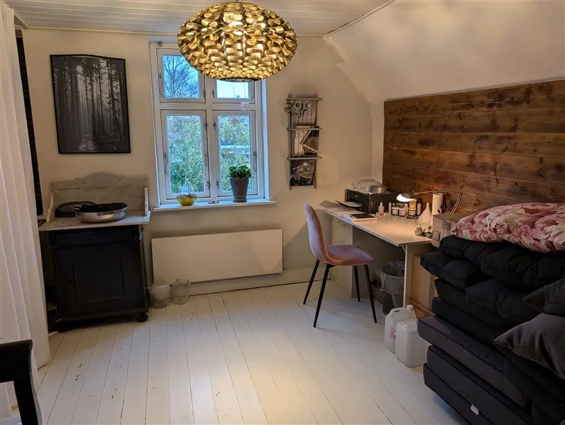 Et lyst værelse med hvide gulvplanker og højt loft med et gulvtæppe, lys gulvlampe, skrivebord, plante og vindue med udsigt