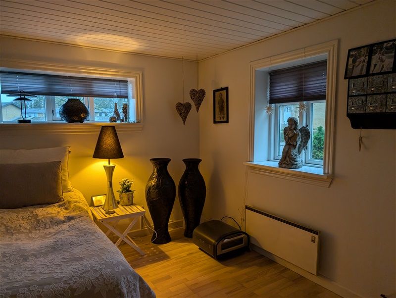 Et soveværelse med dobbeltseng og hvide vægge, trægulv og et sidebord med en lampe og en vase