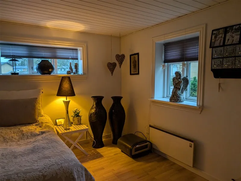 Et soveværelse med dobbeltseng og hvide vægge, trægulv og et sidebord med en lampe og en vase