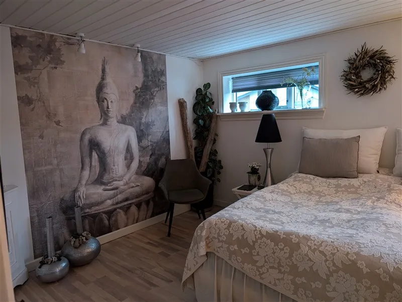 Hjemmets værelse med beige tæppe og hvid pude, Buddha maleri på væggen, lænestol og plante og lysestage på højbordet