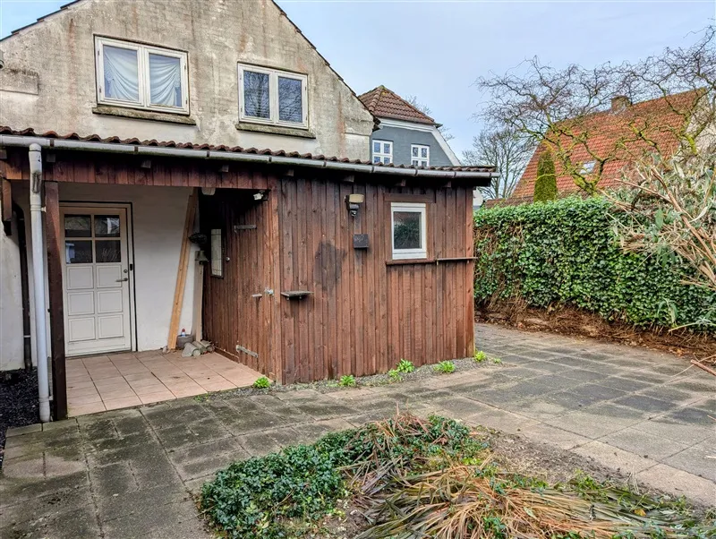 En udstyret træhytte med lyseruder og dør placeret på en haveplads med grønne planter og buske i baggrunden