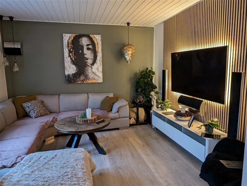 Interiør med hvid sofa og træbord, stor tv-skærm og kunstværk på væggen, lyse pendelaar og lysepunkter under tv-skærmen