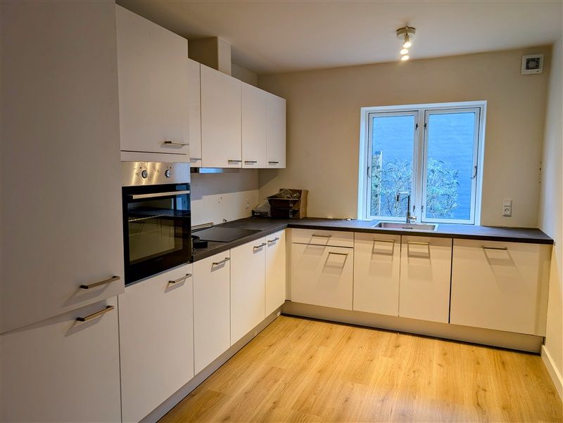 Lys, moderne køkken med hvide vægge og gulve, hvide køkkenskabe og møbler, sort ovn og komfur, lysekroner, vinduer og en