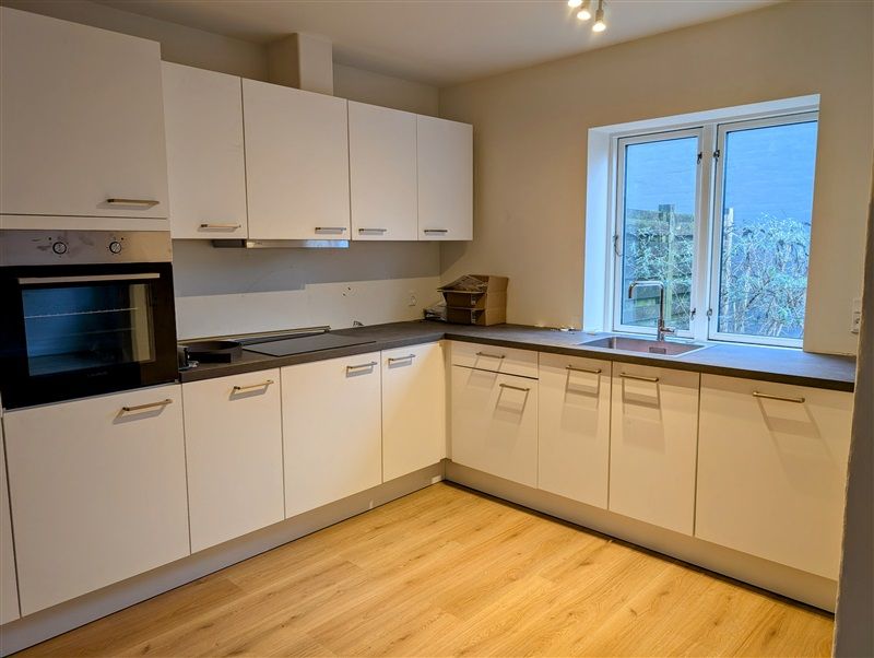 Køkken med hvide skabe og trægulv og lyse loftslamper samt vinduer med udsigt til have