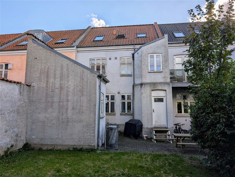 Foto af en bygning med terrasse og grønne planter samt to cykler parkeret foran.