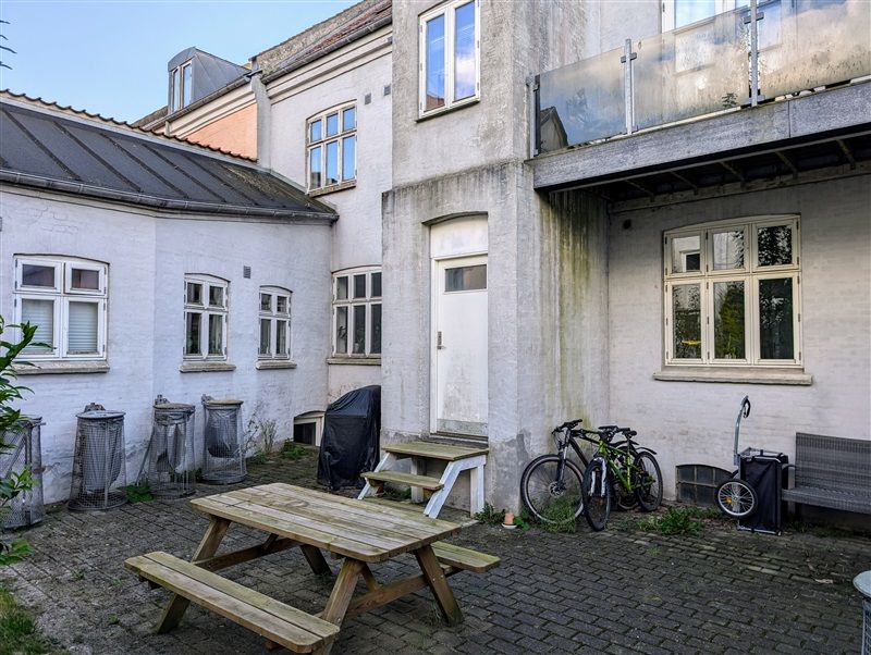 Foto af ejendom med gårdhave og cykler parkeret udenfor. Havebord og bænke er placeret, og der er flere vinduer i baggrunden.
