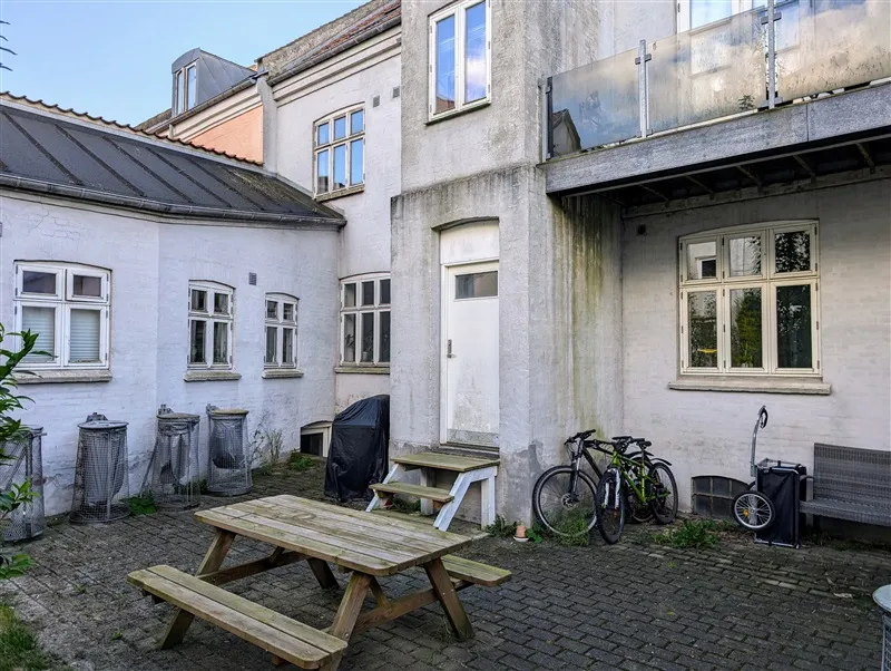 Foto af ejendom med gårdhave og cykler parkeret udenfor. Havebord og bænke er placeret, og der er flere vinduer i baggrunden.