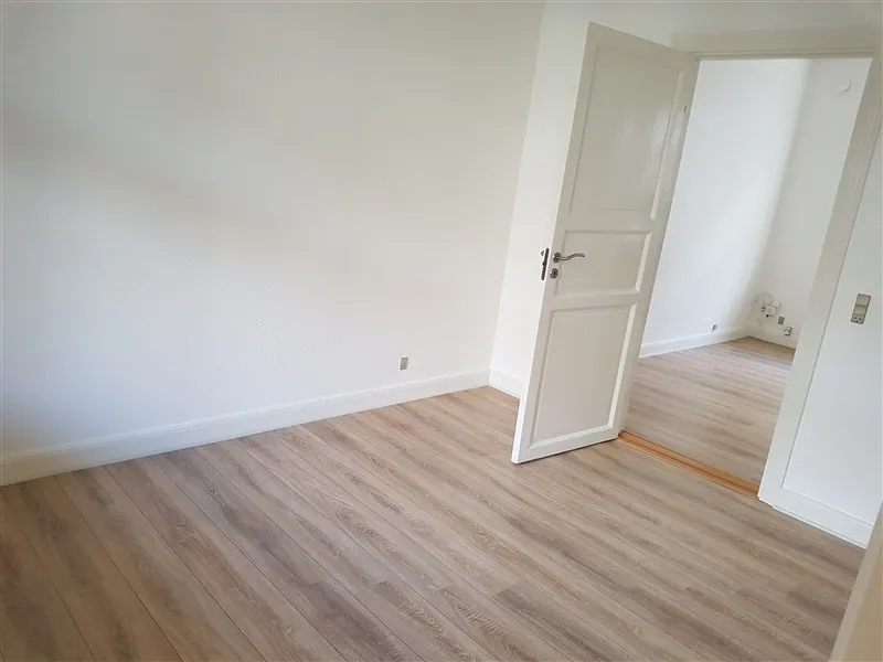 Foto af en stor rummelig lejlighed med hvide vægge og lyse trægulve. Lejligheden har en dør og en åben dør med lyse møbler