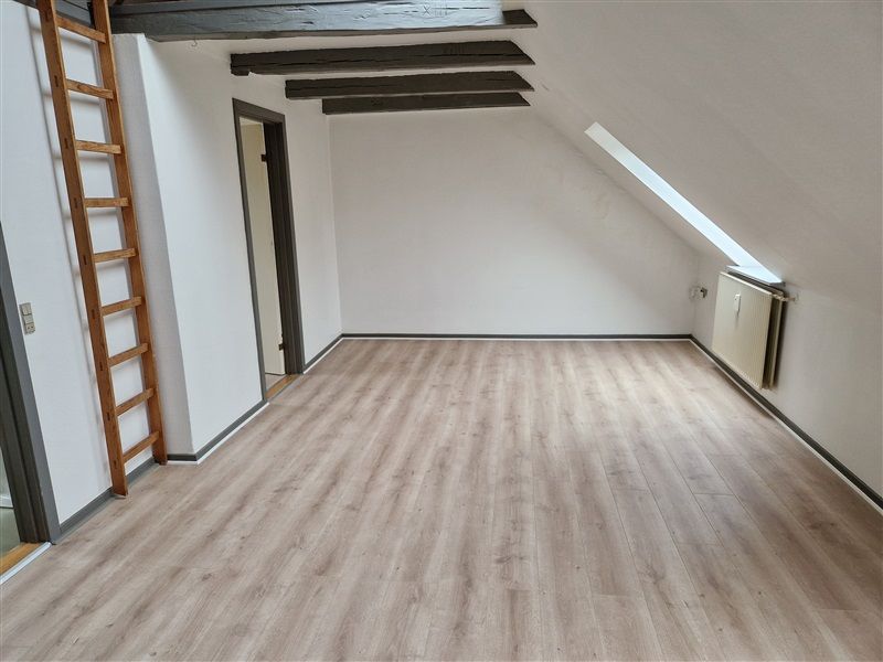 Et tomt rum med hvide vægge, trægulv og loftsbjælker. Der er en trætrappe mod væggen og et radiator på den højre side.