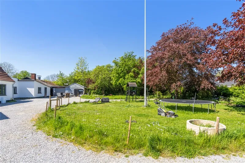 Skur med trampolin og legeplads på græsplads ved siden af hus og vej, lyst og solrigt vejr