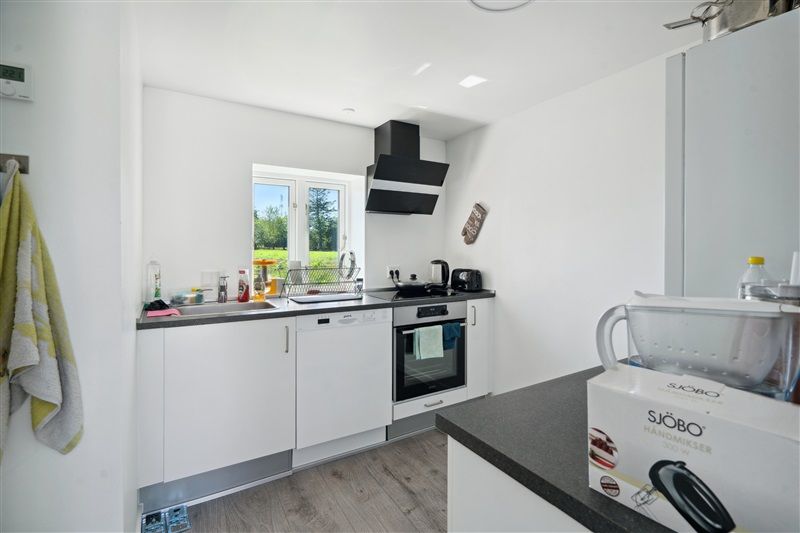 Køkken med hvidt køkkeninventar og lyse loftslampe. En søjleovn og et køleskab er placeret mod væggen. Der er et vindue med