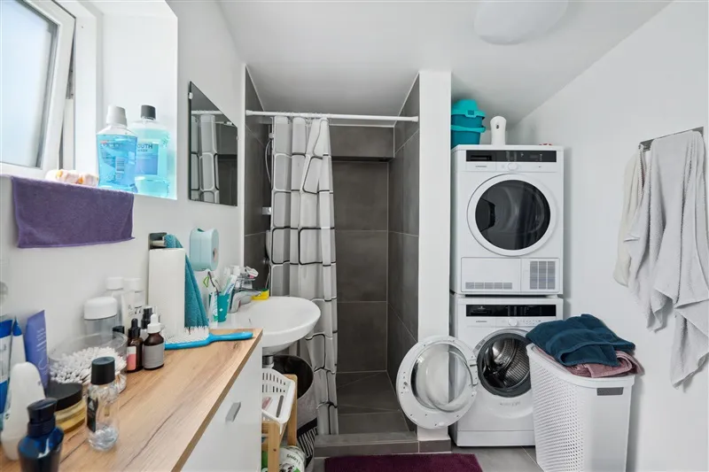 En moderne vaskerum med en hvid maskine og tørretumbler samt et spejl og rengøringsmidler.