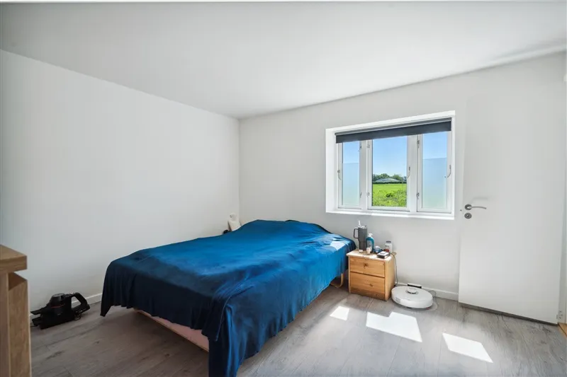 Soveværelse med hvide vægge og gulv, enkeltseng med blå dyne, træskuffe med toiletartikler, vinduesvisker og en søvnig robot.