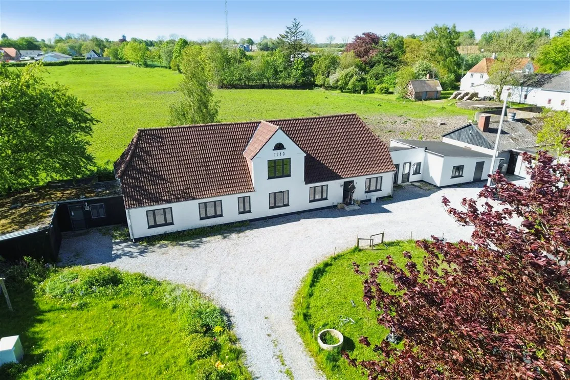 En stor hvid bygning med rødt tag ligger i et landligt område med grønne marker og træer