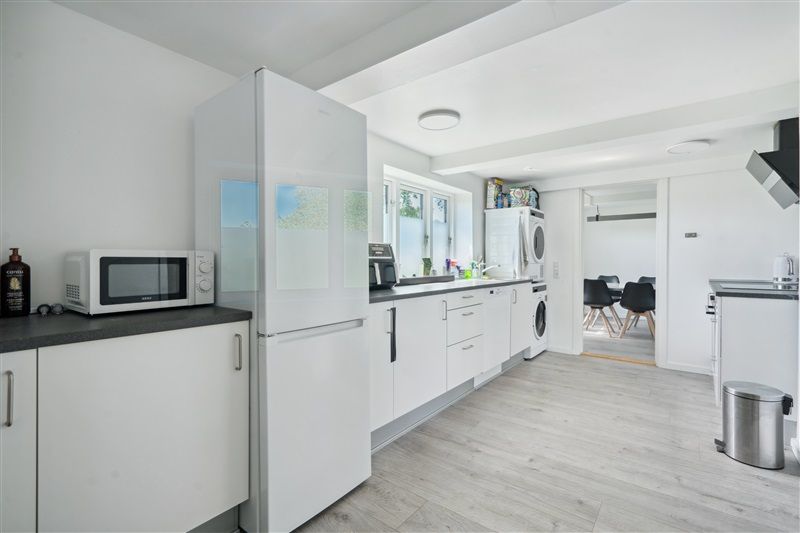 Moderne hvidt køkken med trægulv, hvide skabe, køleskab, mikroovn og vaskemaskine og opvaskemaskine samt en dør til et andet