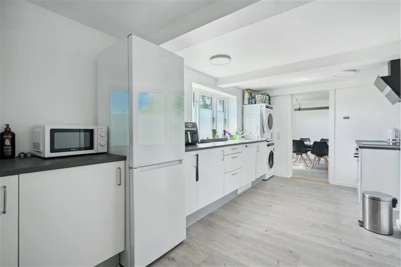 Moderne hvidt køkken med trægulv, hvide skabe, køleskab, mikroovn og vaskemaskine og opvaskemaskine samt en dør til et andet