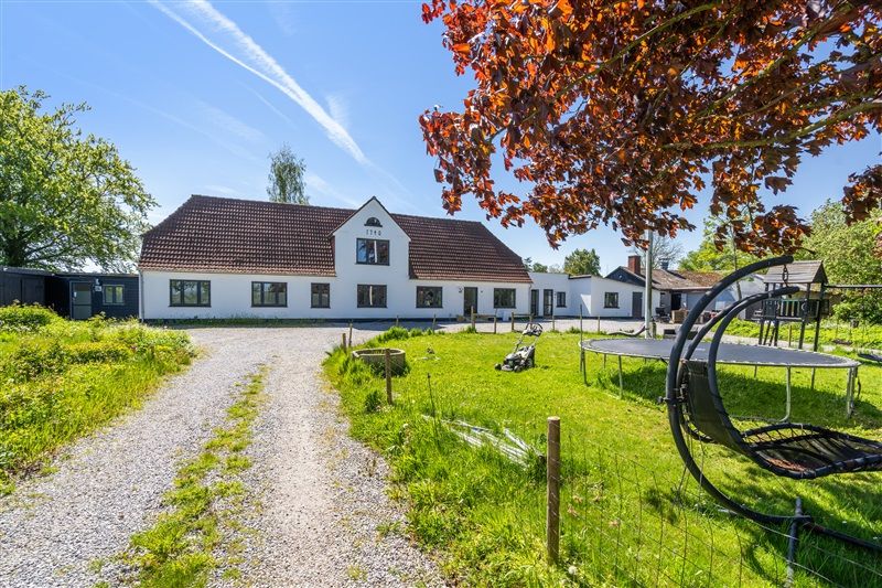 Et stort hvidt hus med brun tagdækning og markerede vinduer, omgivet af en stor græsplæne og en vej, der fører til indkørslen