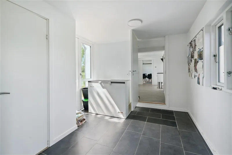 Hallway med hvide vægge og gulv med mørkegrå fliser, spiskammer og åben dør med lyse møbler og gulvtæppe