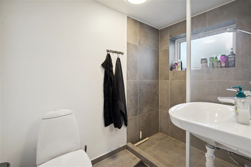 Badrum med hvid toilet, hvid vask og to hængede håndklæder i et moderne interiør med grå mønstre og vinduesglas