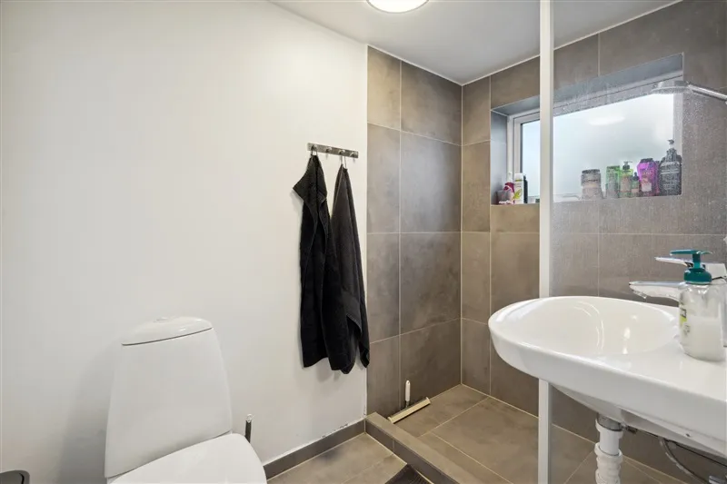 Badrum med hvid toilet, hvid vask og to hængede håndklæder i et moderne interiør med grå mønstre og vinduesglas