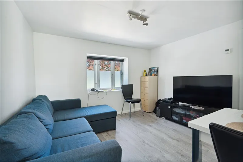 Inde i stuen med hvidt loft og gult gulv er der en blå sofa og en sort stol ved siden af en tv-skærm og et højt skab