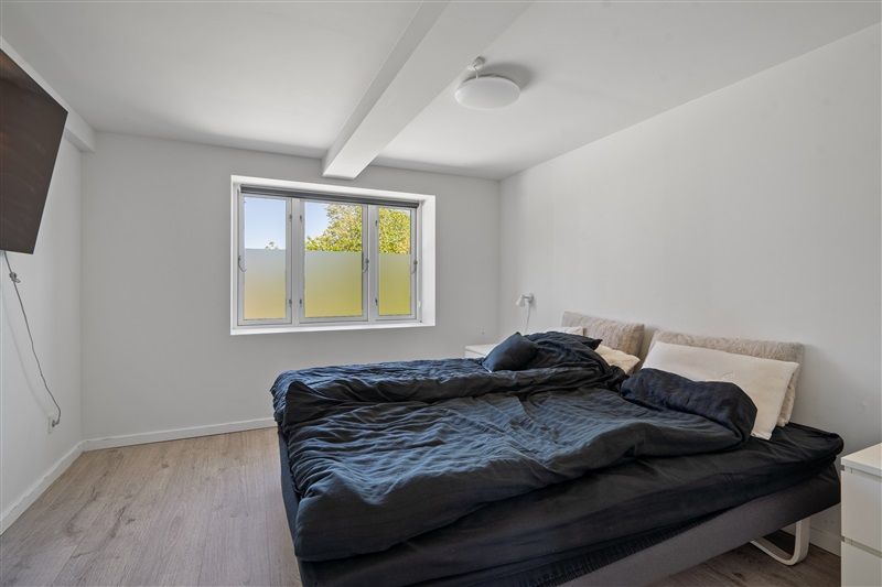 Et moderne soveværelse med en stor seng og hvide vægge og loft. Vinduet har dobbeltruder og giver en dejlig udsigt til haven