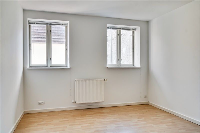 En tom rum med hvide vægge og et trægulv, to store vinduer og en radiator monteret på væggen