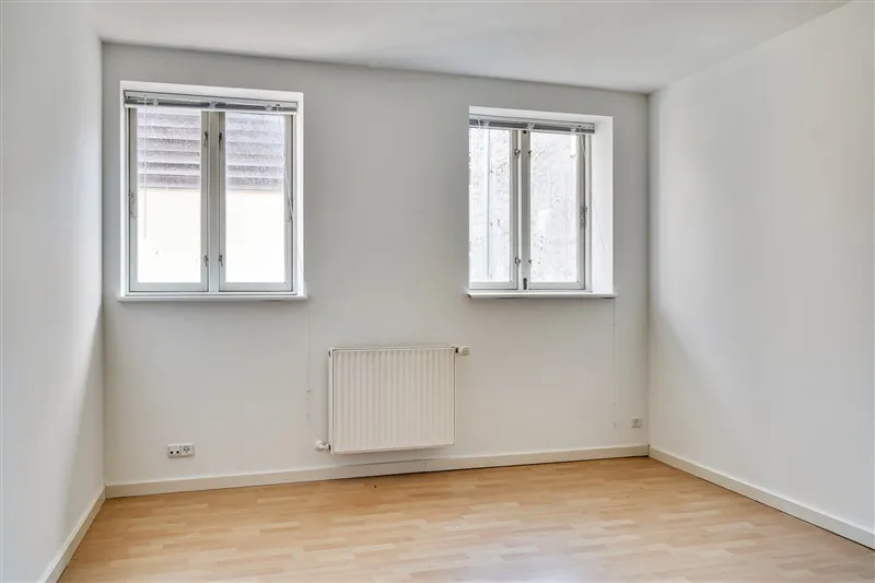 En tom rum med hvide vægge og et trægulv, to store vinduer og en radiator monteret på væggen