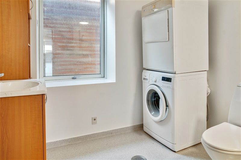 Et lille badeværelse med vask, toilet og en tøjvask og tørretumbler samt et vindue med udsigt til en bygning