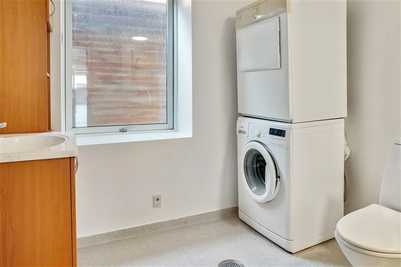 Et lille badeværelse med vask, toilet og en tøjvask og tørretumbler samt et vindue med udsigt til en bygning