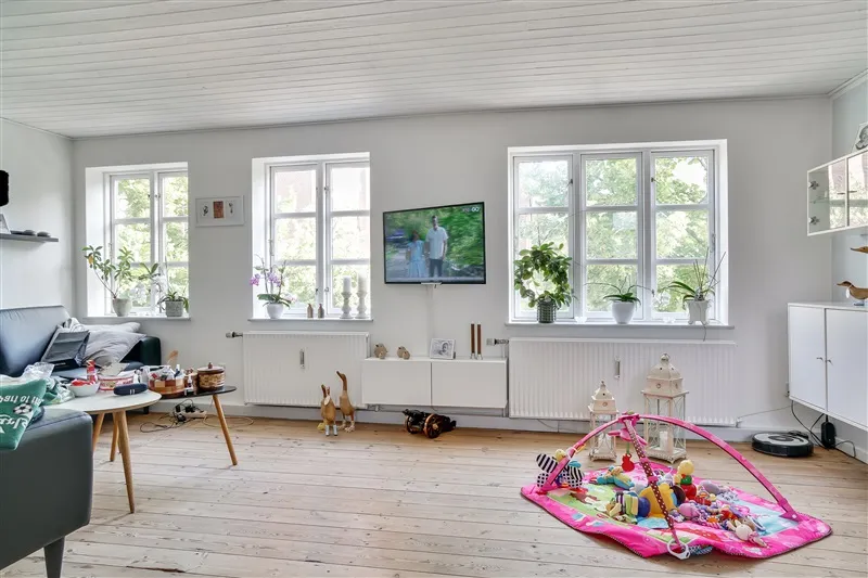 Stue med to smukke hvide vinduer, tv og hvidt gulv, og en sød babylegeplads med legetøj og en pink støtte.