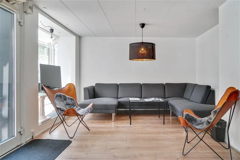 Elegant stue med høj lofthøjde, hjørnesofa i gråt og to orange lænestol med pels. Lampe i mørk metalfarve hænger over bordet