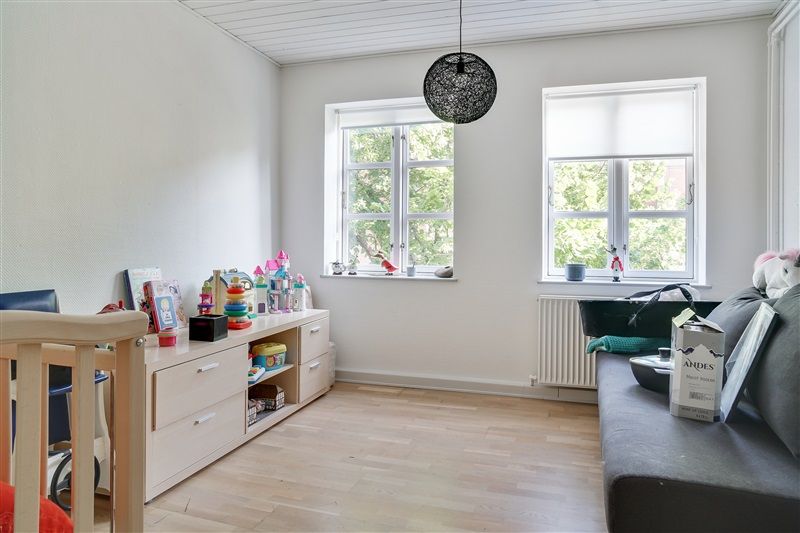 Kig på det lyse værelse med trægulvet, hvide vægge, vindue med gardiner, høj lampe og hylde med legetøj og bøger