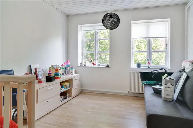 Kig på det lyse værelse med trægulvet, hvide vægge, vindue med gardiner, høj lampe og hylde med legetøj og bøger